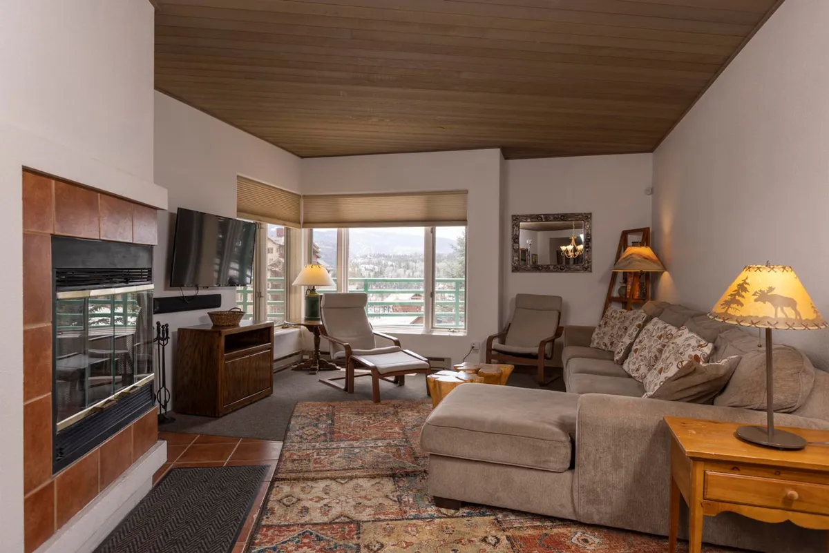 The hero image showcases a cozy living area with a crackling fireplace, a plush sectional sofa accented with patterned pillows, and expansive corner windows revealing breathtaking panoramic views of the Needles Mountains and surrounding ski slopes. The inviting space features warm lighting from themed lamps on wooden side tables, creating a tranquil ambiance perfect for relaxing after a day of adventure.