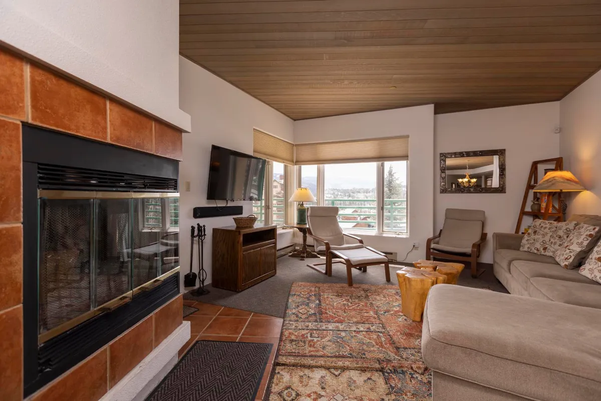 This image showcases a cozy living room anchored by a fireplace, with a comfortable sectional sofa, large windows offering scenic mountain views, and a wooden lounge chair with ottoman. The setup is inviting and perfect for gathering or unwinding after a day of mountain adventures.