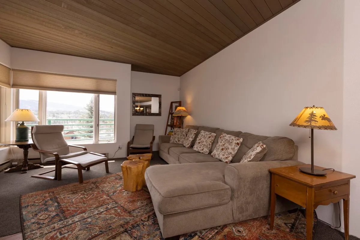 A spacious interior scene featuring a large sectional sofa with patterned cushions, wooden accent chairs near a window, and decorative tree-themed lamps. The arrangement faces large windows that frame panoramic mountain vistas, emphasizing the airy, open feel of the living space.