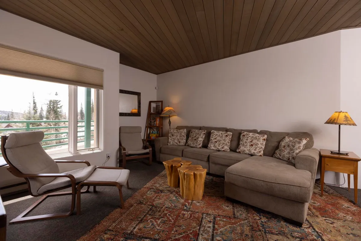 A cozy living room with a beige sectional sofa, rustic wooden stump coffee tables, two cushioned armchairs by a window, and a wood-paneled ceiling. The window invites natural light and offers glimpses of the mountain landscape, reinforcing the space’s serene, rustic atmosphere.