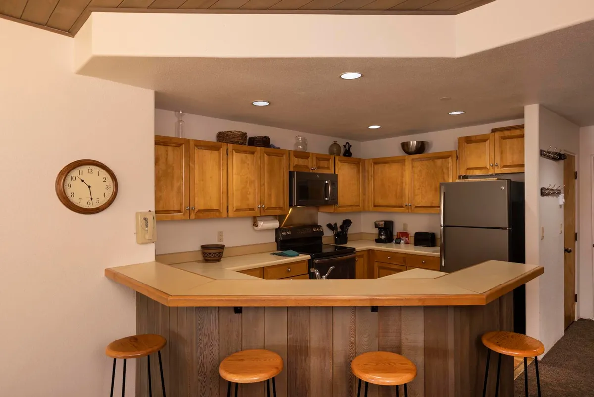 The kitchen space is showcased with rustic wooden cabinets, a hexagonal breakfast bar with stools, and modern stainless steel appliances, including a refrigerator and microwave. The wall clock adds a homey touch, and the bar area invites casual interaction and quick meals.
