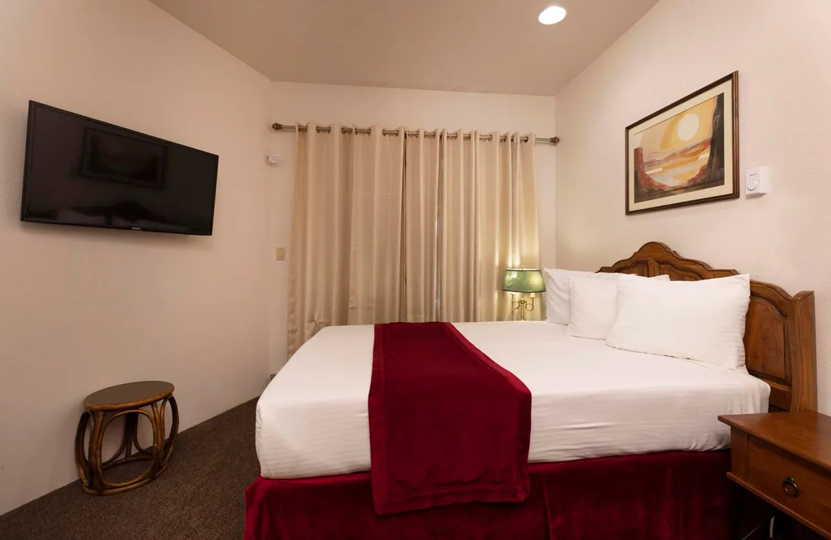This image displays a serene bedroom with white linens and red accents, a wall-mounted flat screen TV, framed artwork above the bed, and curtains covering the window. The warm, inviting atmosphere suggests a cozy space where guests can relax after a day of mountain adventures.