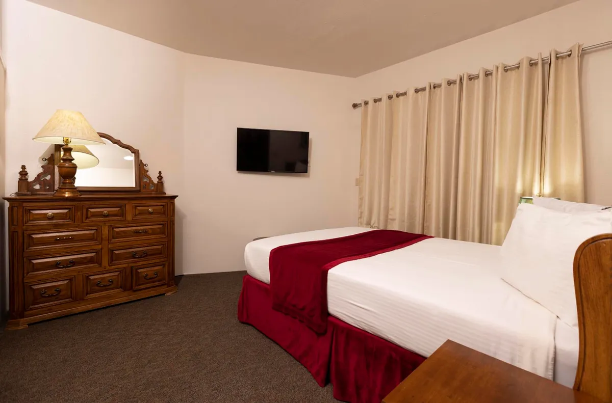 A serene bedroom scene with a bed dressed in crisp white linens and a bold red blanket, complemented by a wooden dresser, a mirror, and a soft-glow lamp. A wall-mounted flat-screen TV and thick curtains add to the sense of privacy and relaxation, making this room a perfect place to unwind after mountain adventures.