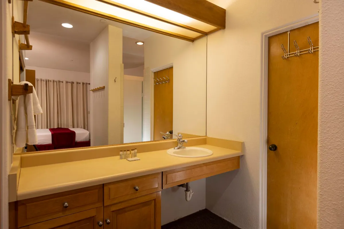 This image shows a spacious bathroom with a wide countertop, large mirror reflecting the bedroom, wooden cabinetry, and a door with hooks for towels or robes. The calm, practical setting hints at a private space for rejuvenation and self-care.