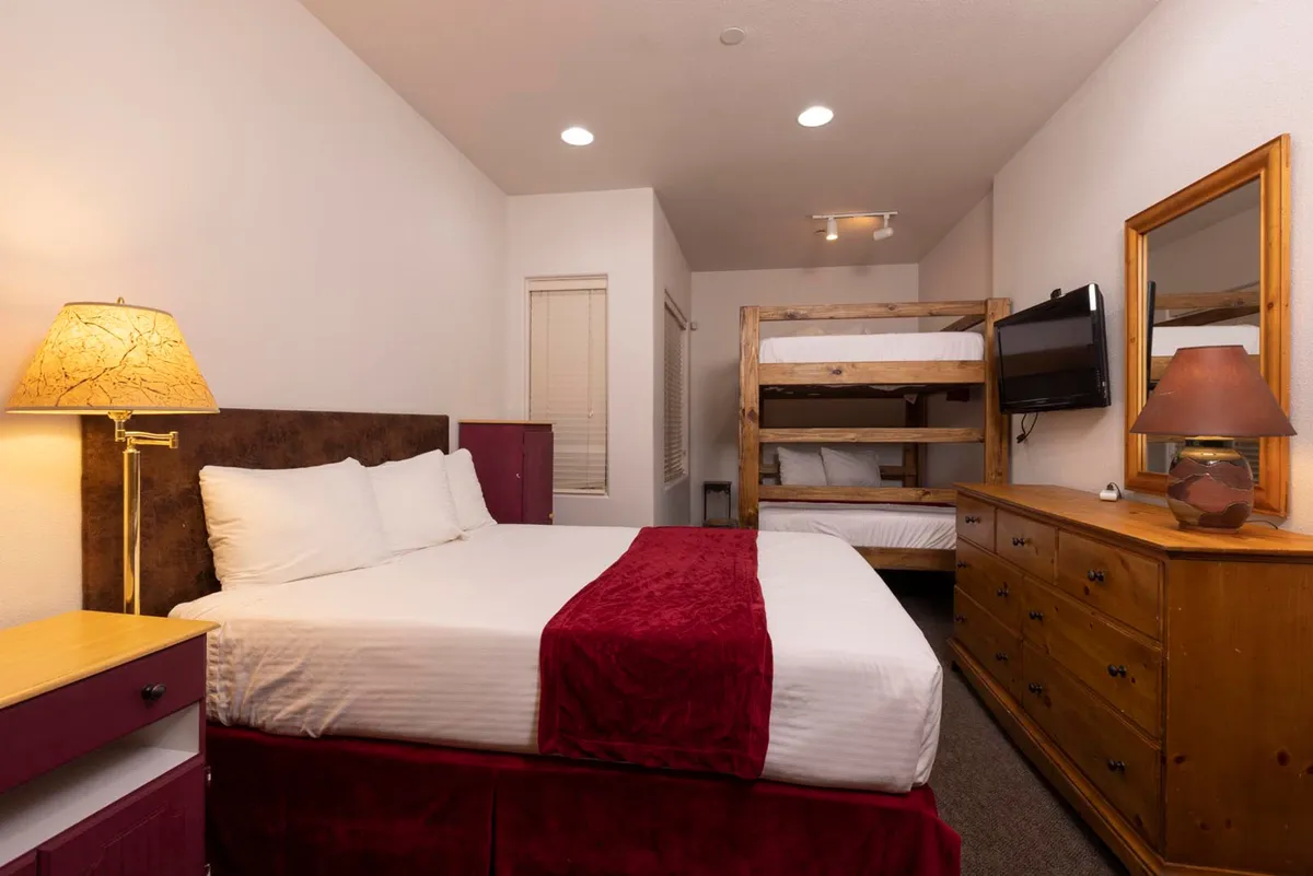 A cozy, inviting bedroom with a large bed draped in a red blanket, wooden bunk beds, a dresser with a mirror and lamp, and a mounted flat screen TV. The warm lighting and practical layout suggest comfort and relaxation after an active day outdoors.