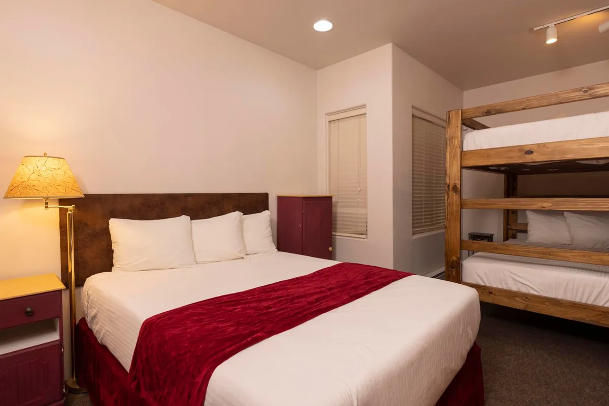 A spacious, inviting bedroom featuring a king-sized bed adorned with a red runner, a practical wooden bunk bed for extra guests or children, a warm bedside lamp, and convenient storage with a purple bedside table and cabinet. The room is softly lit, creating a cozy and welcoming space ideal for families or groups seeking comfort after a day outdoors.