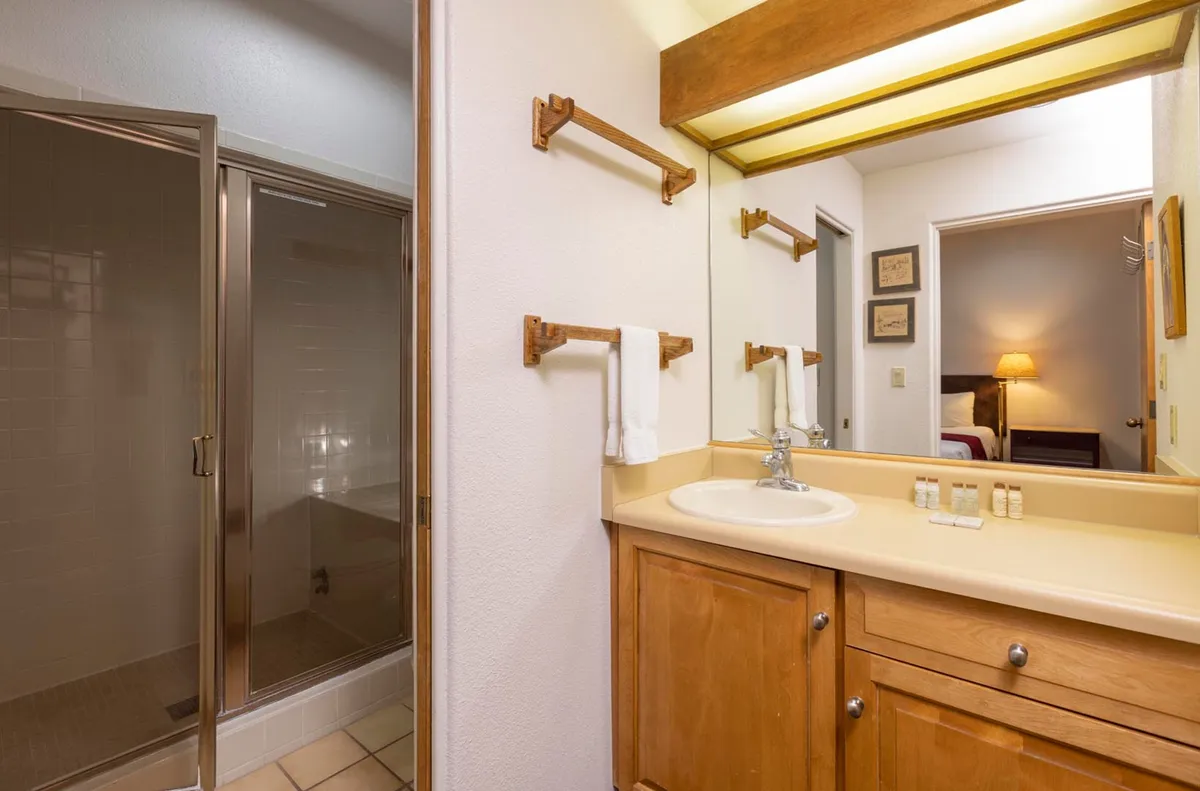 A clean, well-lit bathroom featuring a wood-framed mirror with overhead lighting, a wooden vanity with a beige countertop and sink, glass-enclosed shower stall, and neatly arranged white towels. The space feels inviting and practical, supporting both energizing morning routines and relaxing evening soaks.