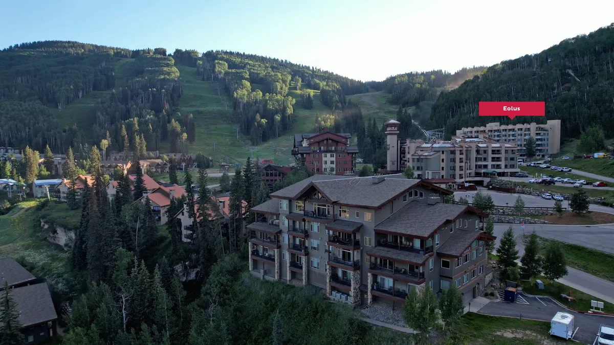 A wide exterior shot featuring the main resort building with multiple balconies, surrounded by pine trees and natural landscape. The ski slopes can be seen on the forested mountainside behind the resort, and the building labeled 'Eolus' is visible, situating the viewer at the heart of the resort. The image hints at numerous on-site amenities, including access to the heated pool and hot tubs.