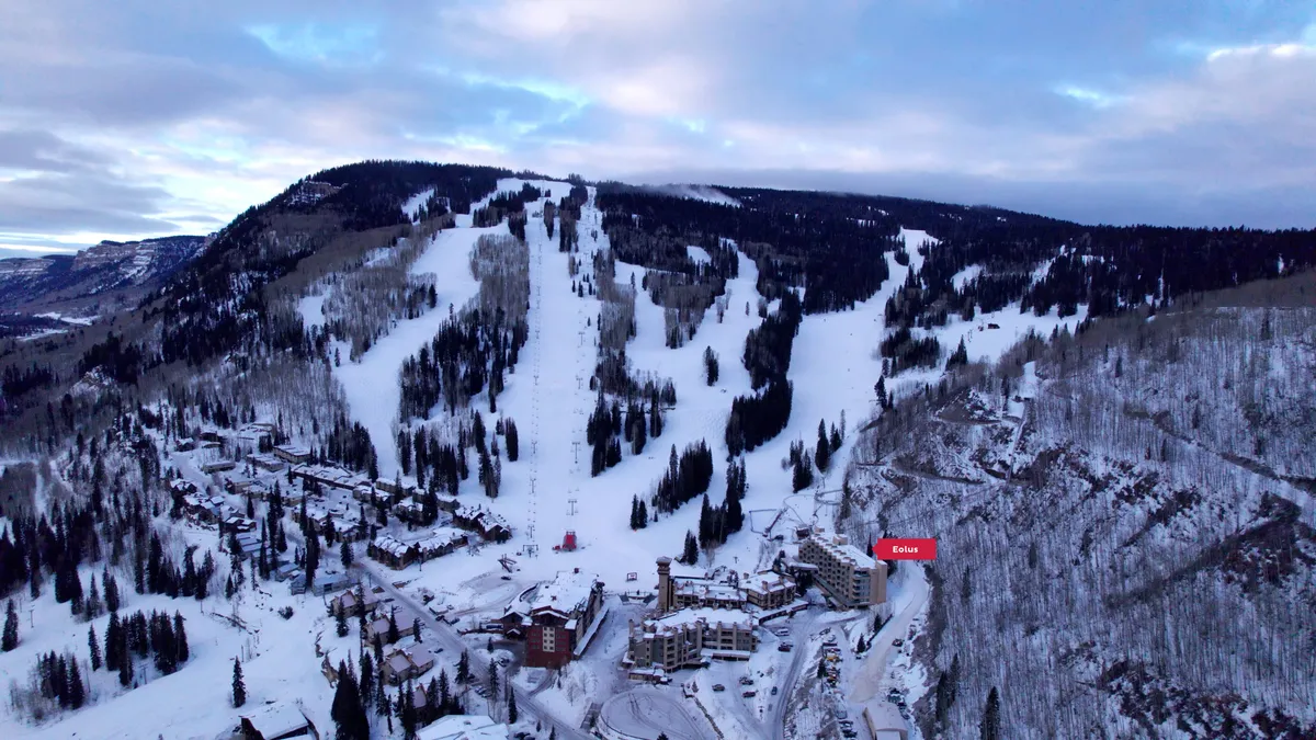 A sweeping exterior view of Purgatory Resort's snow-covered slopes, ski lifts, and surrounding pine and aspen forests. Multiple resort buildings cluster at the mountain base, with winding roads and pathways connecting them. The dramatic Needles Mountains are visible in the background, creating a sense of adventure and immersion in nature.