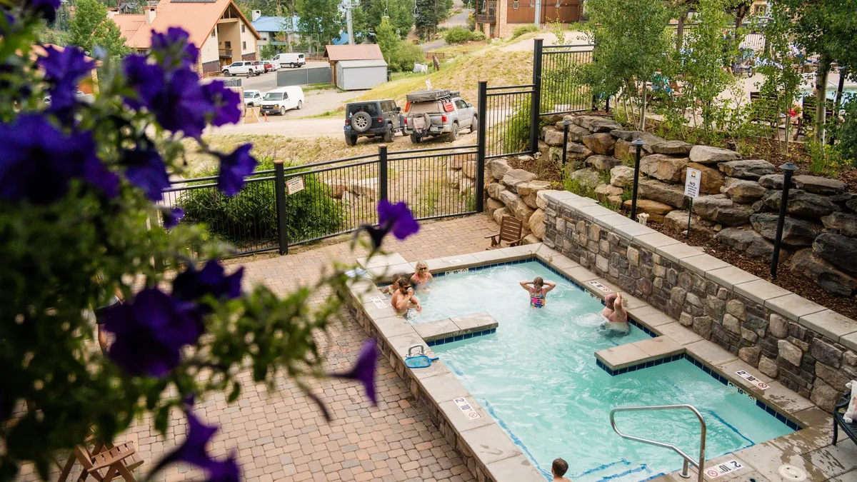 This exterior image captures the heated community pool in use, with people swimming and relaxing. The pool is framed by a stone wall, with vibrant purple flowers in the foreground, and resort buildings and vehicles visible in the background.