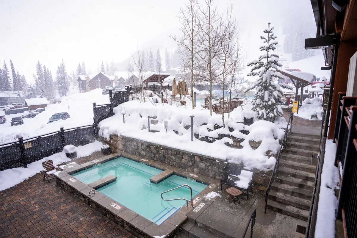 This exterior shot highlights a heated outdoor pool with steam rising into the crisp winter air, surrounded by snow-laden trees and stone walls. Patio furniture and distant rooftops further set a tranquil winter resort scene.