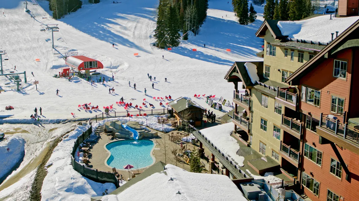An animated exterior scene with skiers on the slopes, a heated outdoor pool with a waterslide, and red lounge chairs set against the snowy landscape. The multi-story resort building with balconies can be seen in the background, emphasizing the seamless blend of adventure, relaxation, and family-oriented amenities.