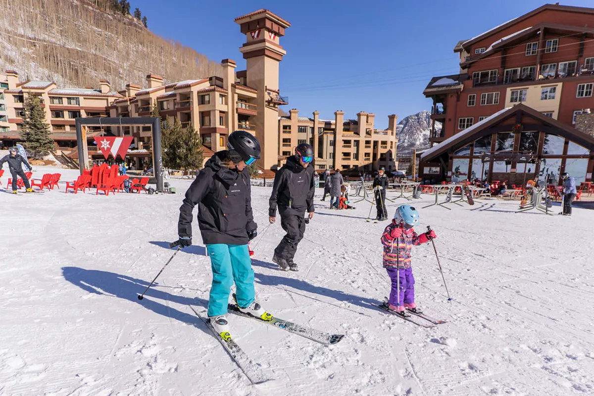 A lively winter scene with skiers of all ages, including a child learning to ski, set against snow-covered ground and resort buildings. The image includes visible ski equipment, an outdoor seating area, and panoramic mountain views.